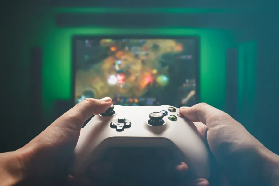 Imagem de uma pessoa segurando um controle branco, jogando videogame. A tela da TV ao fundo mostra o jogo.