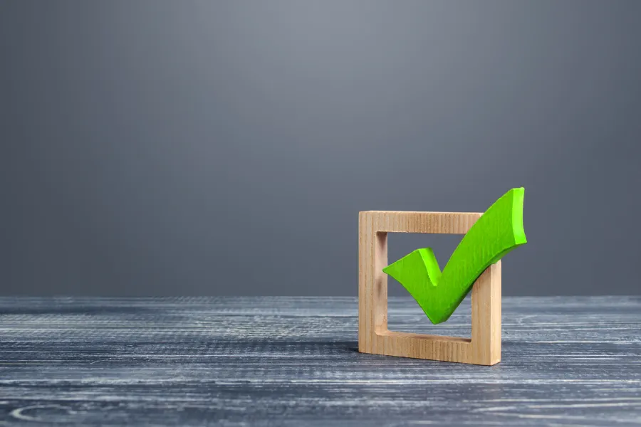 Símbolo de checkmark verde tridimensional em uma caixa de seleção quadrada de madeira, sobre uma superfície escura, representando aprovação, conformidade ou conclusão.