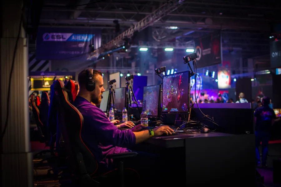 Um jogador de e-sports com fone de ouvido e microfone, concentrado em seu computador durante uma partida competitiva, com luzes de palco e banners de e-sports ao fundo.