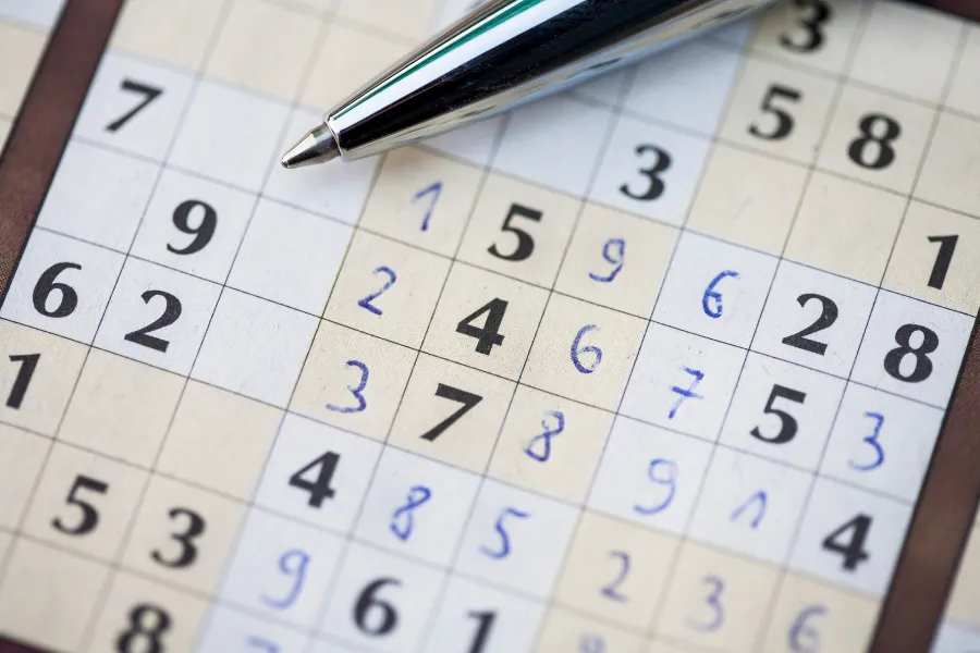 Um close-up de um quebra-cabeça de Sudoku em papel, com a maioria dos quadrados preenchidos com números em tinta azul e um 4 e 7 em negrito, e uma caneta prateada na parte superior.