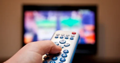 Mão segurando controle remoto de TV com tela colorida desfocada ao fundo.