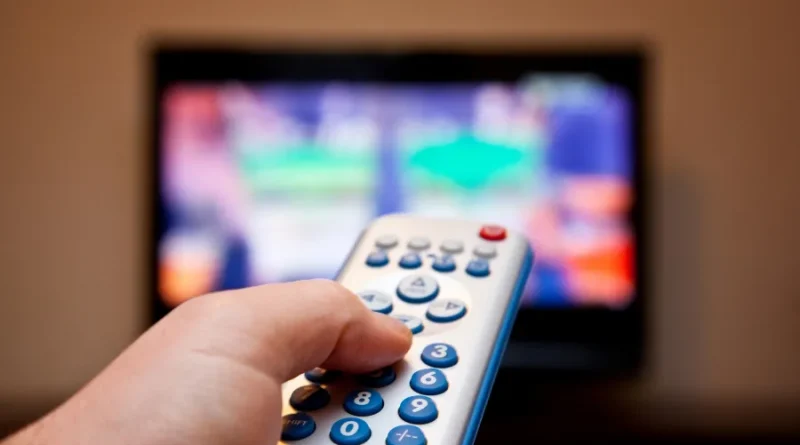 Mão segurando controle remoto de TV com tela colorida desfocada ao fundo.