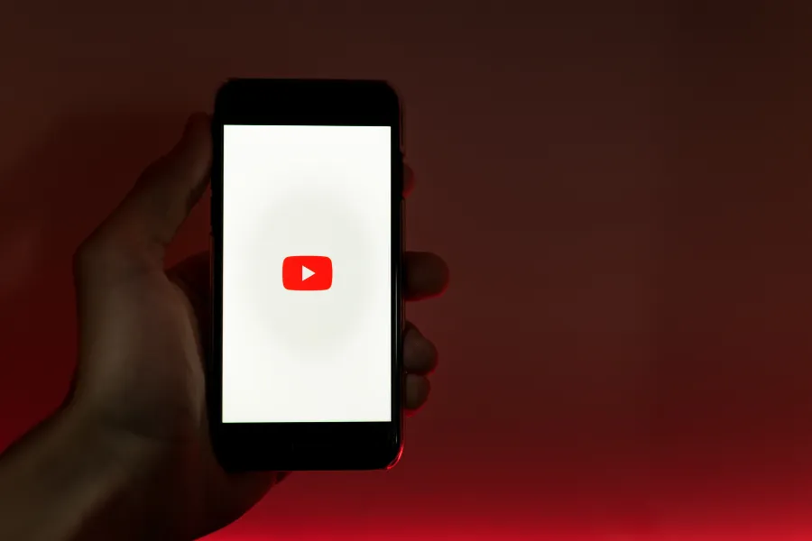Mão segurando smartphone com o logotipo vermelho do YouTube na tela branca.

