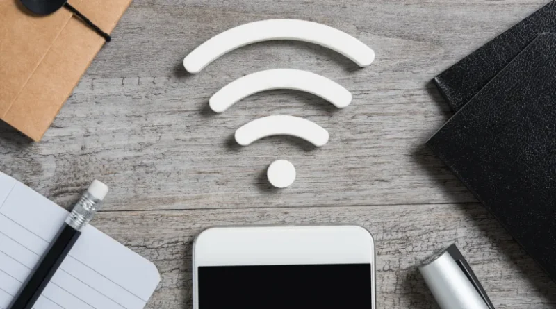 Imagem de um smartphone com um ícone branco de Wi-Fi acima, sobre uma mesa de madeira com objetos como um caderno, lápis e caneta.